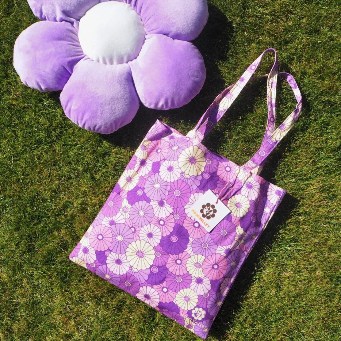 Cotton purple floral patterned tote bag in different shades of purple laying on the grass next to a big flower cushion