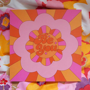 An 8 inch art print with a floral shaped motif with rays beaming from the centre in pink, yellow and orange. The words Be You with three stars  inside the circle.
