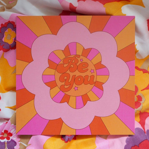 An 8 inch art print with a floral shaped motif with rays beaming from the centre in pink, yellow and orange. The words Be You with three stars  inside the circle.