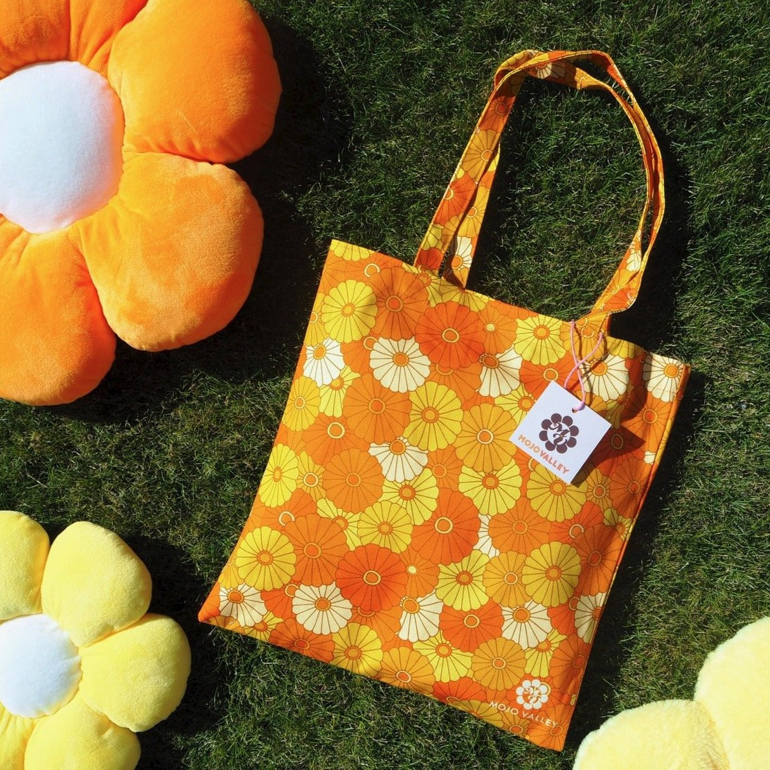 Cotton citrus floral patterned tote bag in different shades of yellow and orange laying on the grass next to two flower cushion