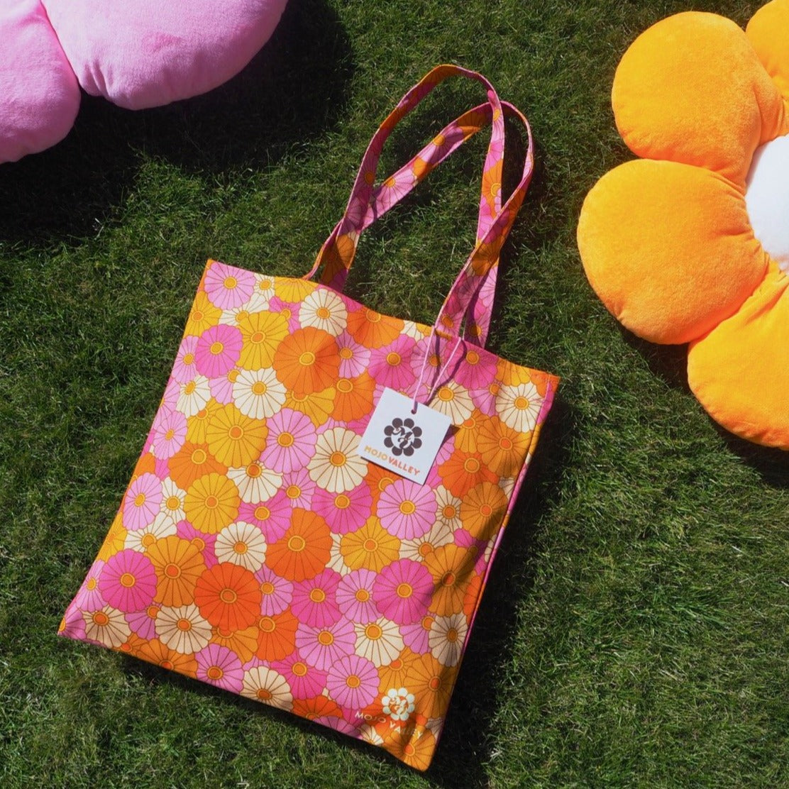 Cotton tutti frutti floral patterned tote bag in pink, yellow, orange and cream laying on the grass next to two big flower cushion